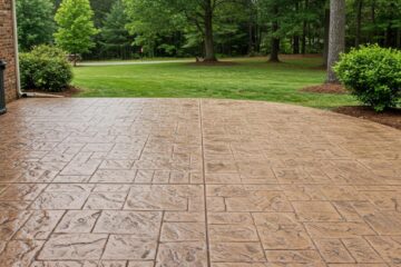 ### Main Subjects
- **Patio Surface**: A textured, stamped concrete patio.
- **Surrounding Nature**: Lush greenery including trees and shrubs.

### Notable Features
- **Color and Texture**: Earthy brown tones with a stone-like pattern on the patio.
- **Landscape**: Open grassy area with trees in the background, creating a natural setting.

### Text or Symbols
- **None Present**: The image does not contain any text or symbols.

### Overall Mood or Theme
The image conveys a serene and inviting atmosphere, highlighting the harmony between the man-made patio and the natural landscape. The wet surface suggests a recent rain, adding freshness to the scene.